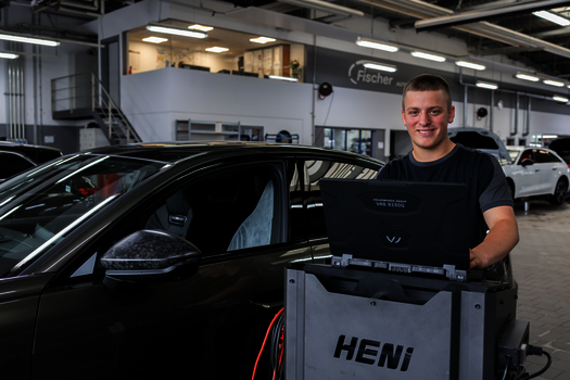 Junger Mann steht in einer Halle neben einen KFZ und bedient ein digitales Messgerät 